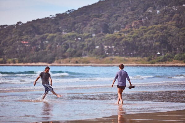 Two people walking on a beach with waves in the background and a forested hill. - Lorne Foreshore Caravan Park (Lorne)