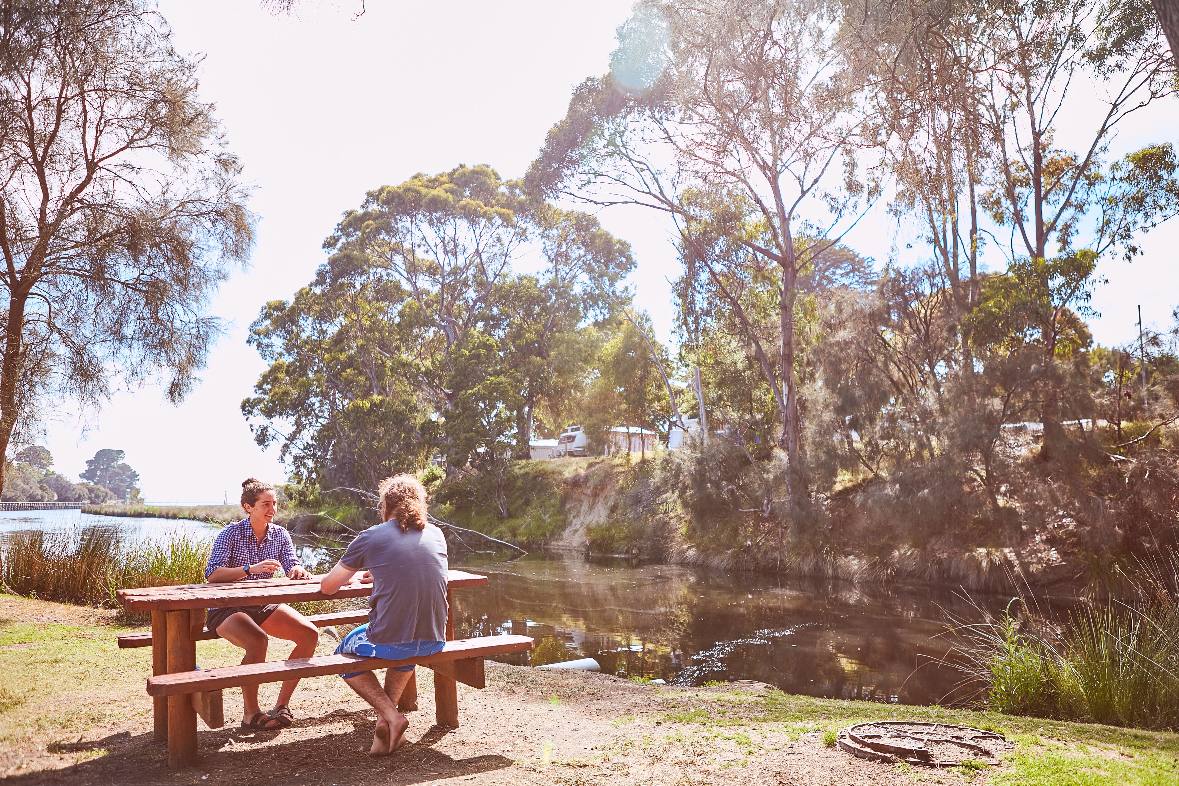A picnic table by a lake with people sitting and one person fishing.