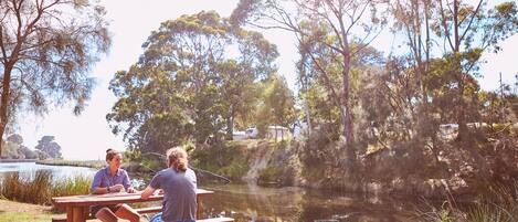 A picnic table by a lake with people sitting and one person fishing.
