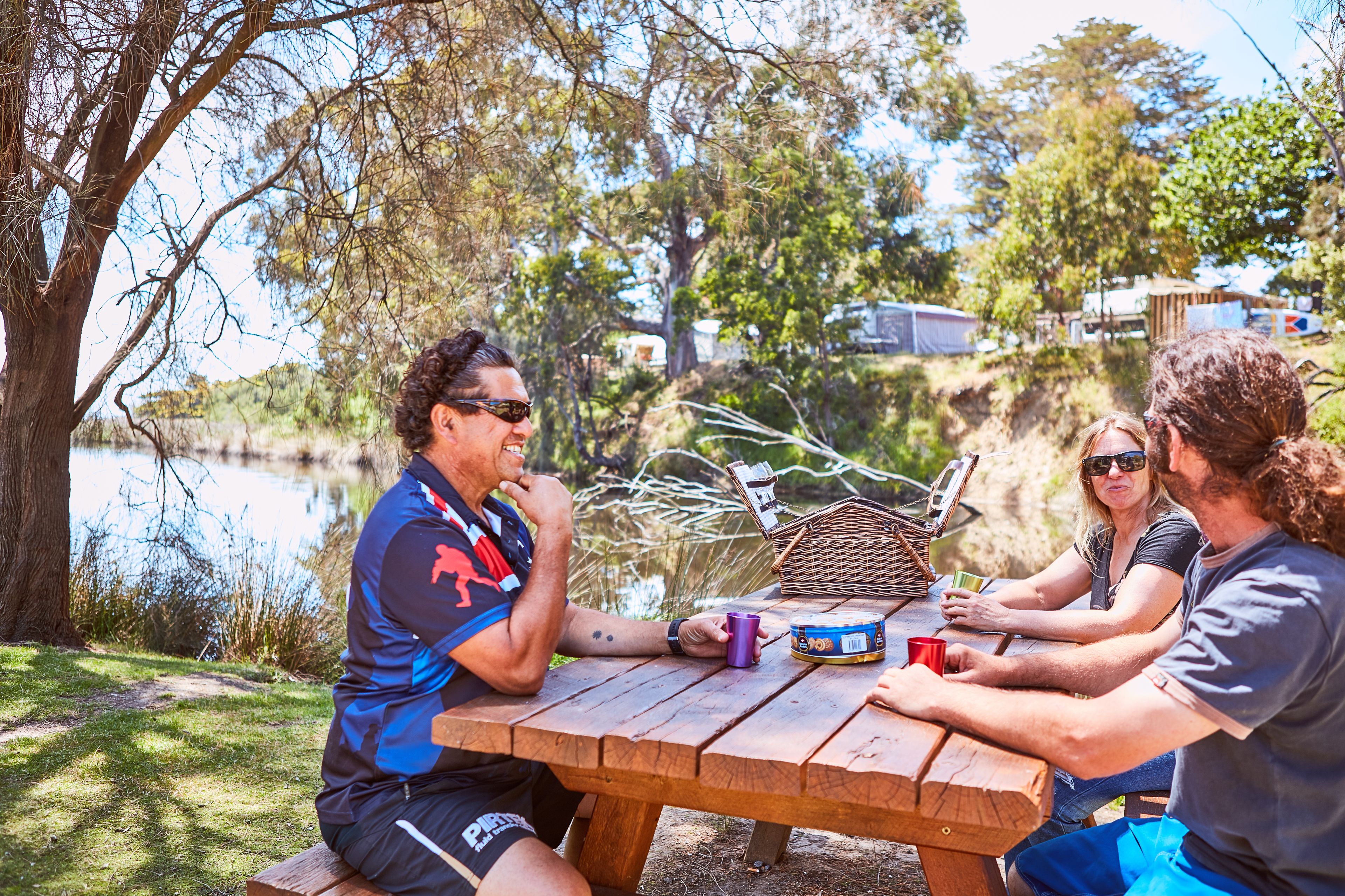 Un grupo de personas sentadas en una mesa de picnic de madera al aire libre, con un lago y árboles de fondo.