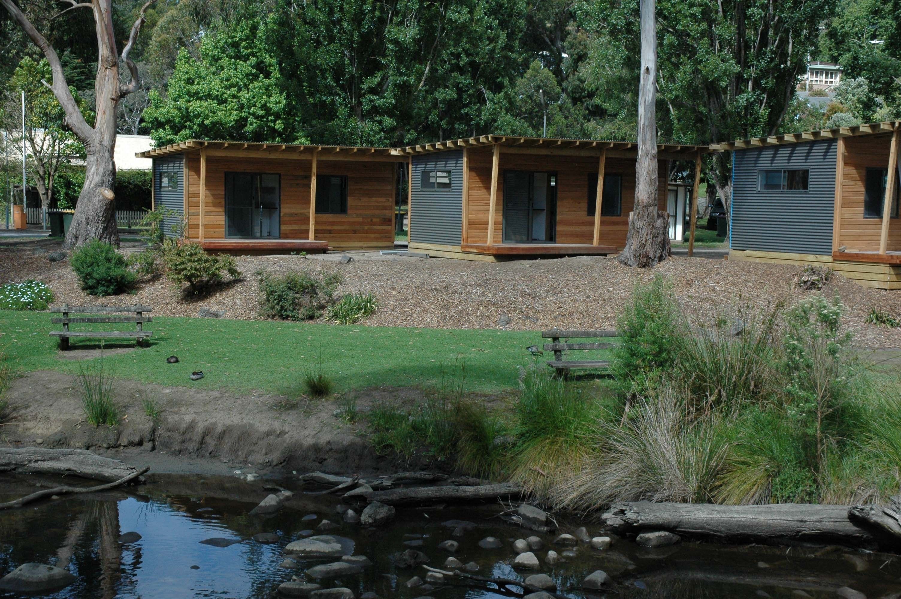A row of wooden cabins with large windows and a small pond in the foreground.