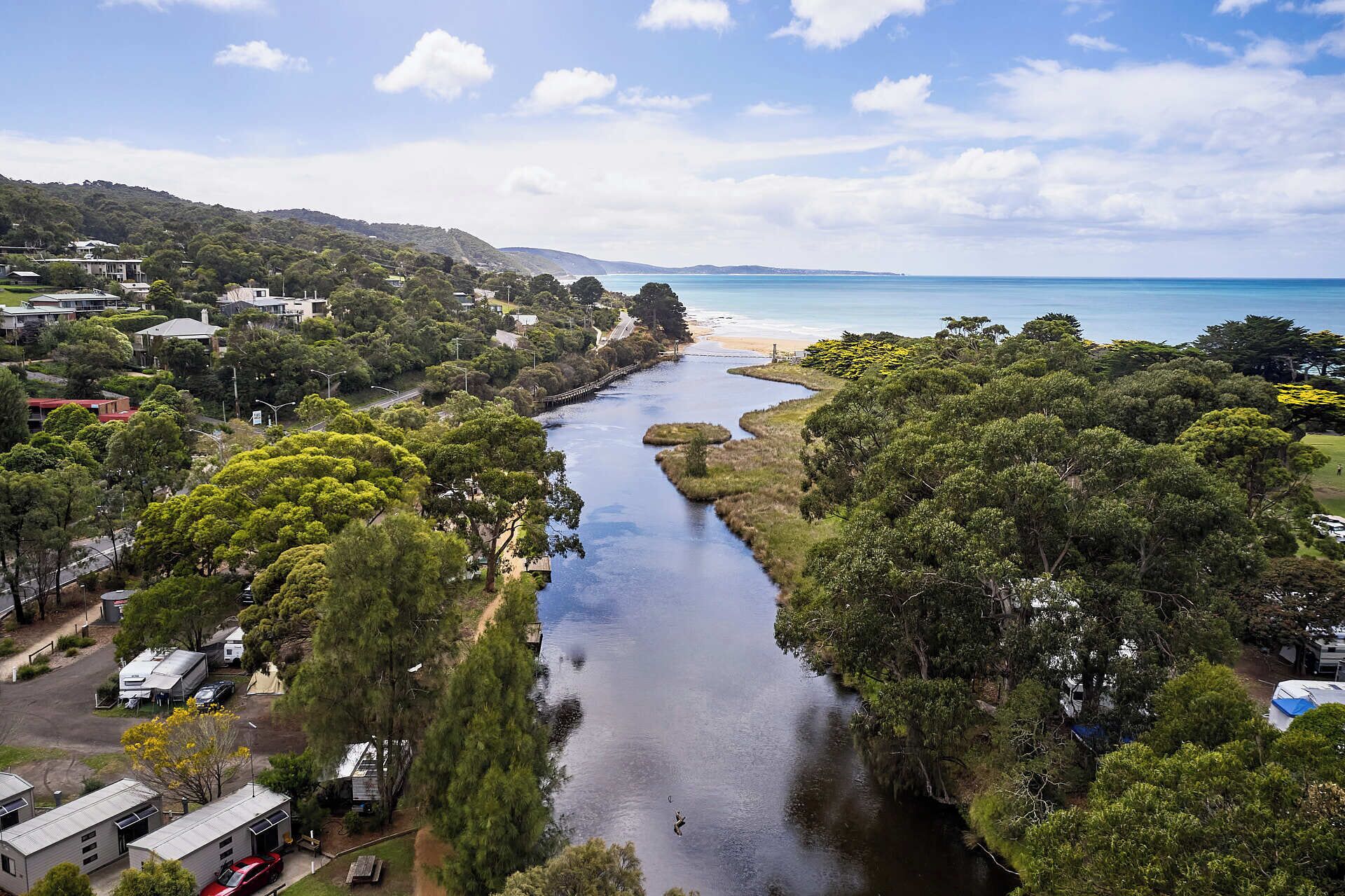 A river flowing through a densely wooded area with a beach and houses in the background.