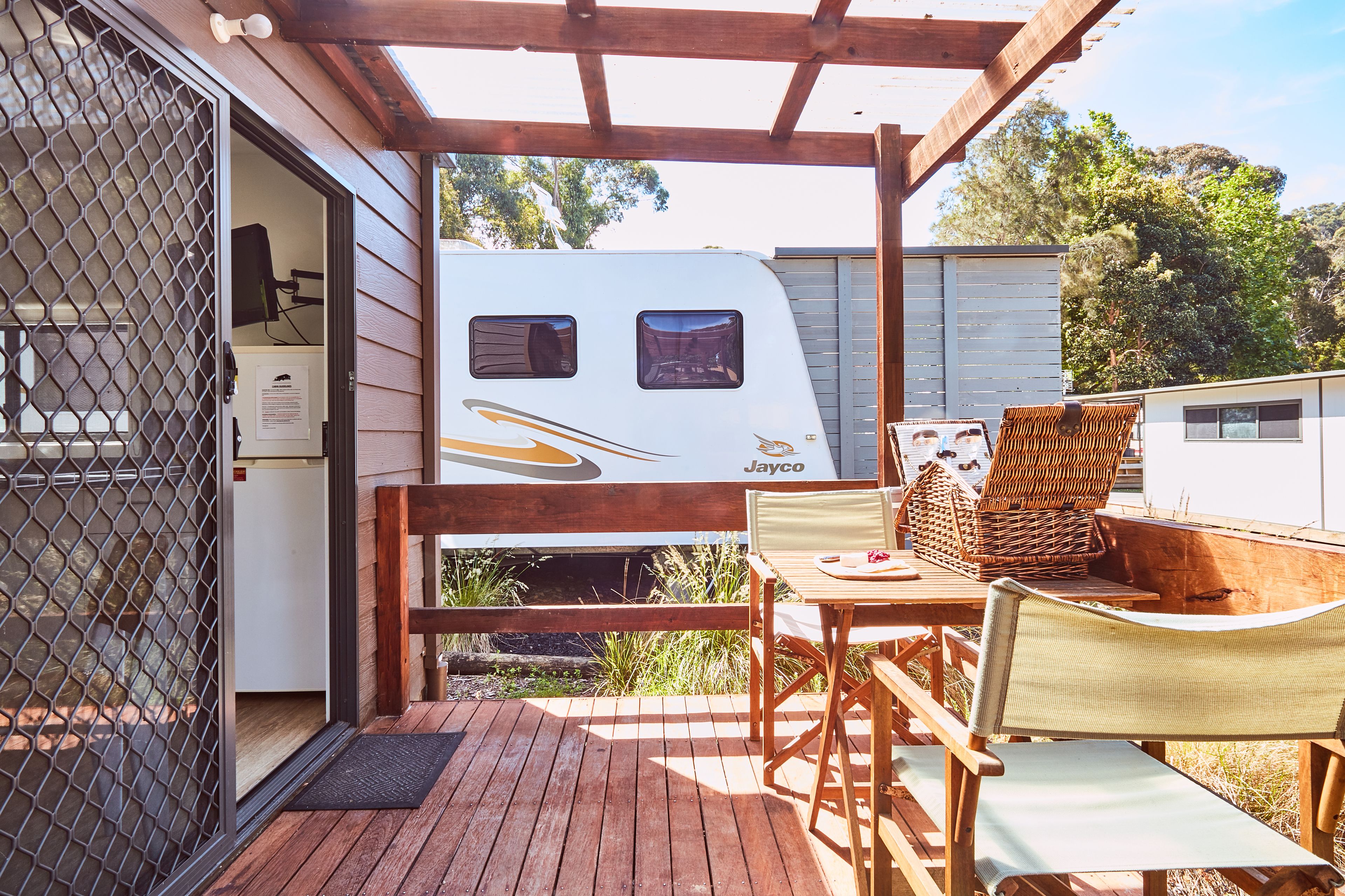 Un espace extérieur abrité, doté d’une terrasse en bois, d’une table avec des chaises, et d’un camping-car blanc stationné derrière une grille métallique.
