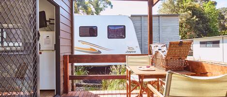 Un espace extérieur abrité, doté d’une terrasse en bois, d’une table avec des chaises, et d’un camping-car blanc stationné derrière une grille métallique.