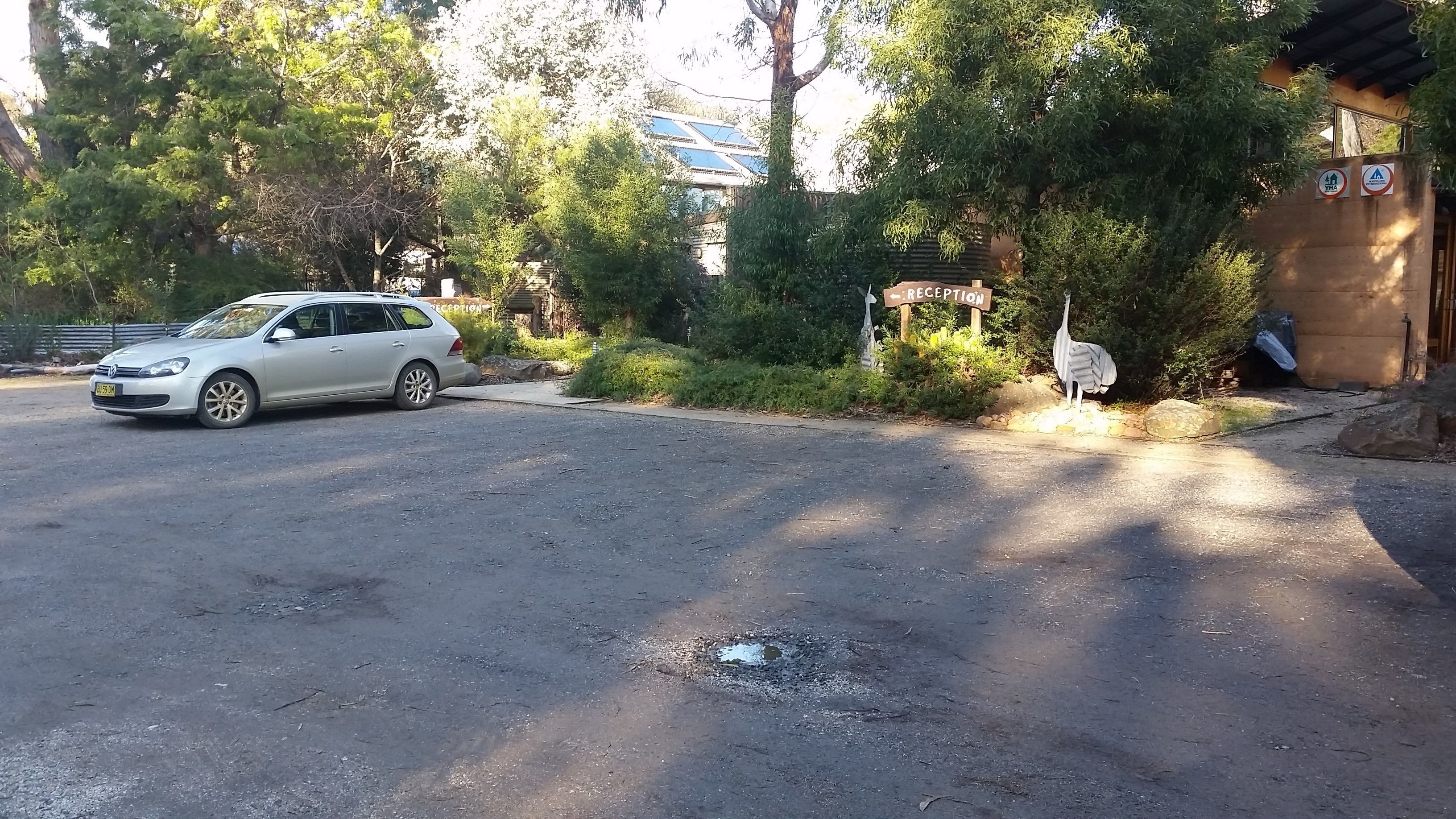 Un coche blanco aparcado en un camino de tierra con un cartel de recepción y una estatua de pájaro cerca.