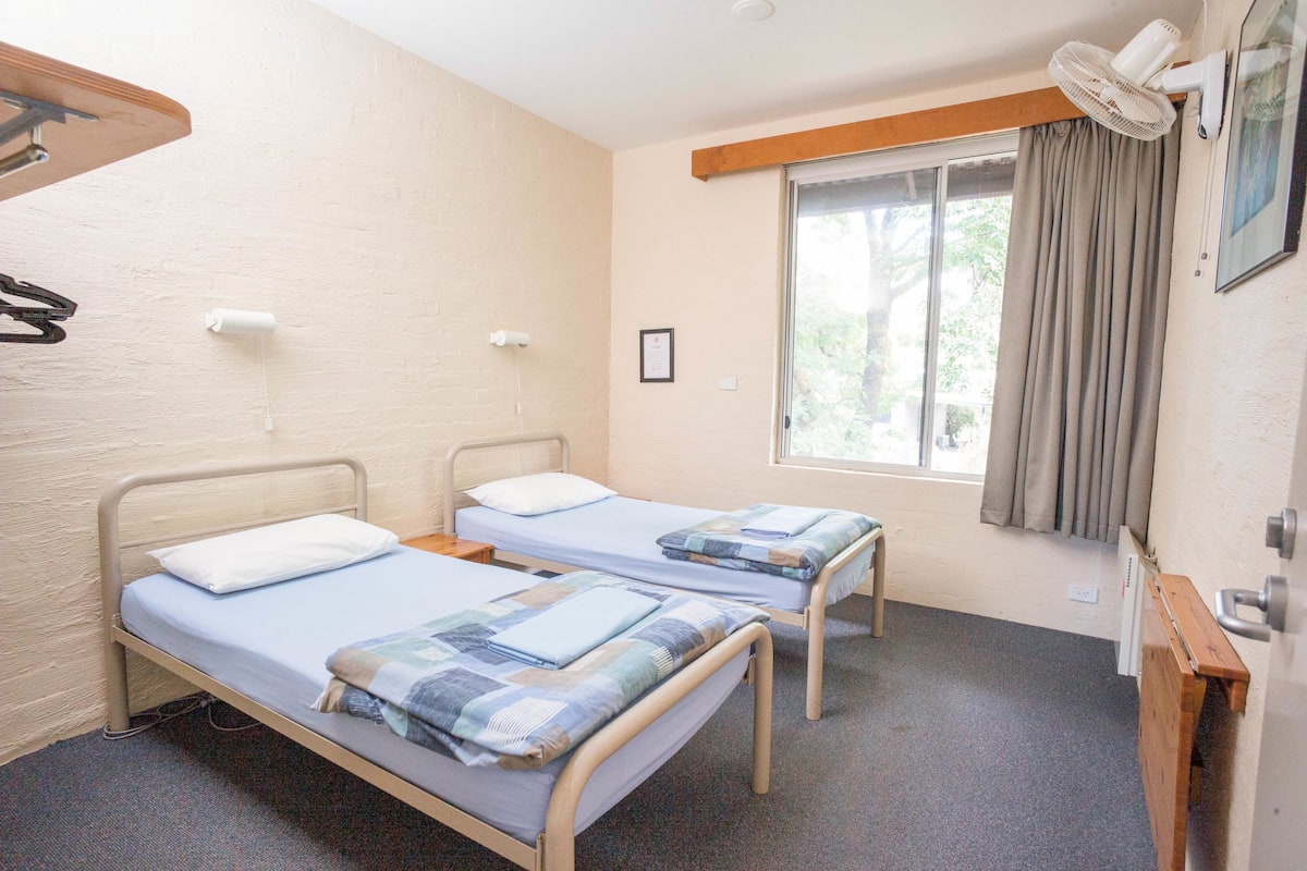 Una habitación con dos camas individuales, una ventana con cortinas, un ventilador de techo y un estante de madera.