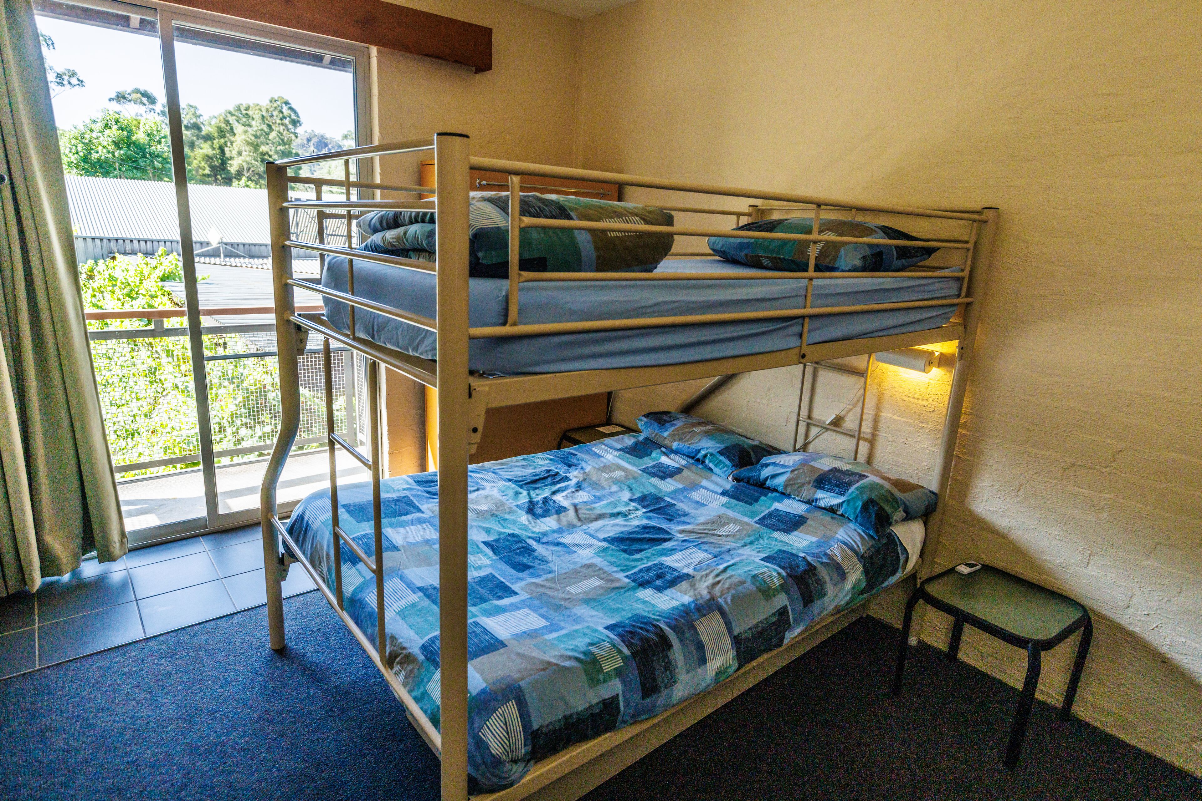A bunk bed with blue bedding, a small table, and a window with a view of greenery.