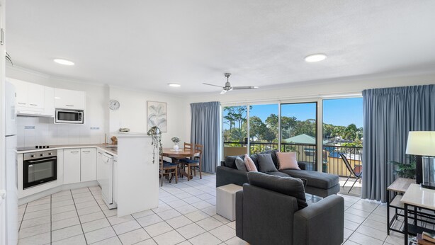 A modern kitchen with white cabinets, a microwave, and an oven. There is a dining area with a table and chairs, and a grey sofa in the living area. - Alexander Beachfront Apartments (Torquay)