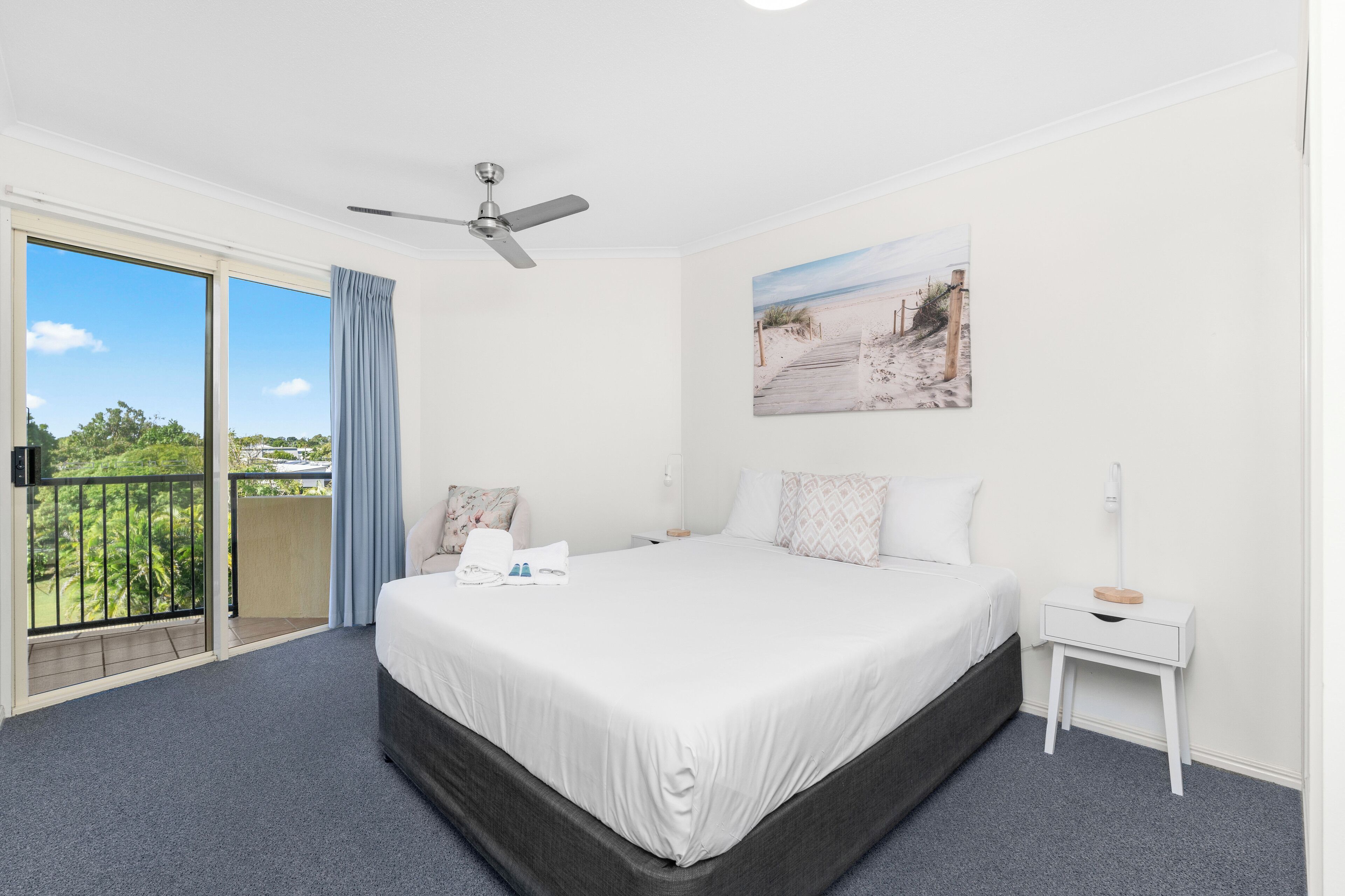 A hotel room with a large bed, a ceiling fan, a painting of a beach, a balcony with a view of greenery, and a white nightstand with a lamp.