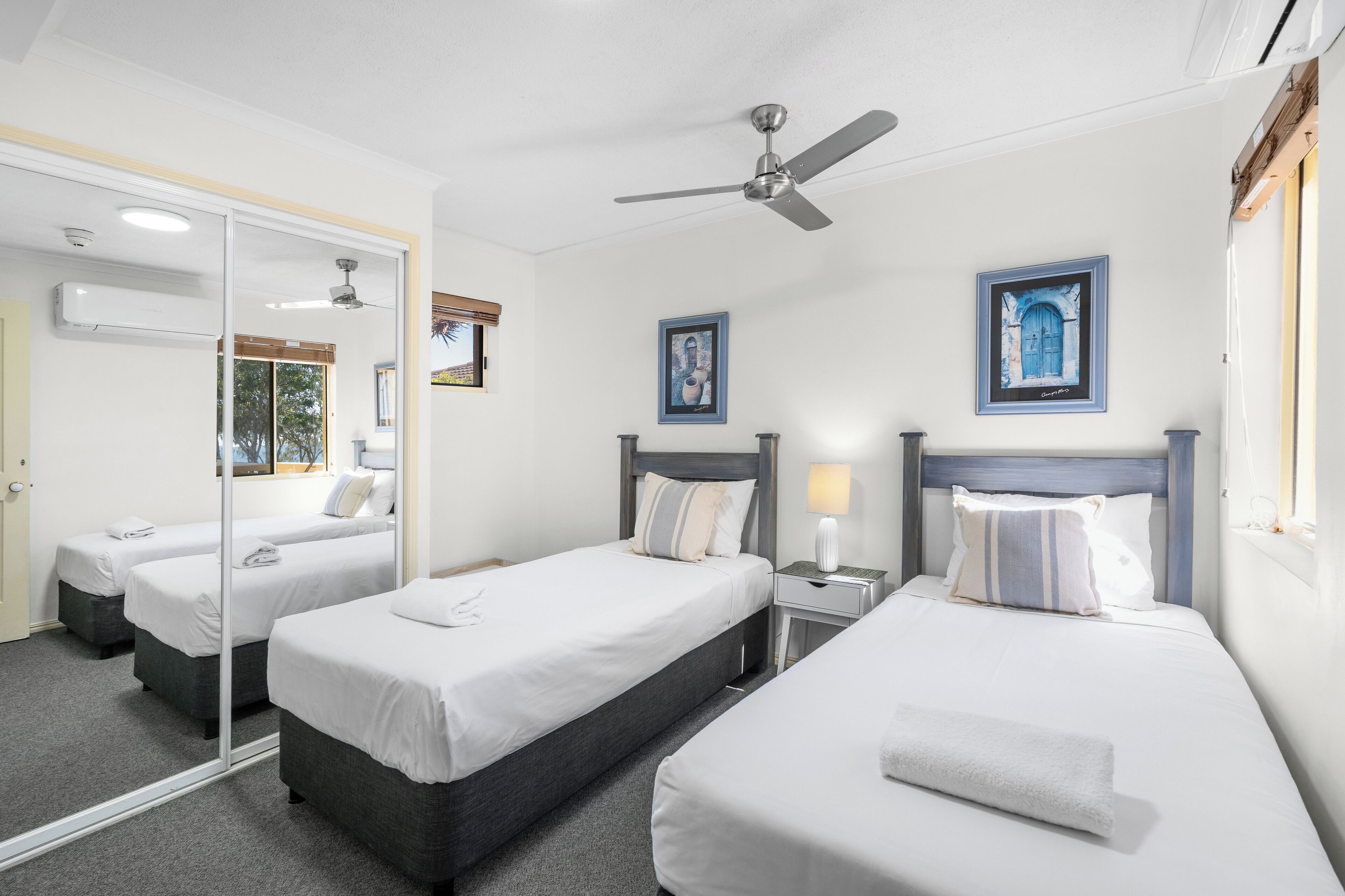 A hotel room with two beds, a ceiling fan, a mirror, and artwork on the walls.
