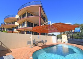 Un edificio de varios pisos con balcones, piscina y una zona con sombra con mesa y sillas.