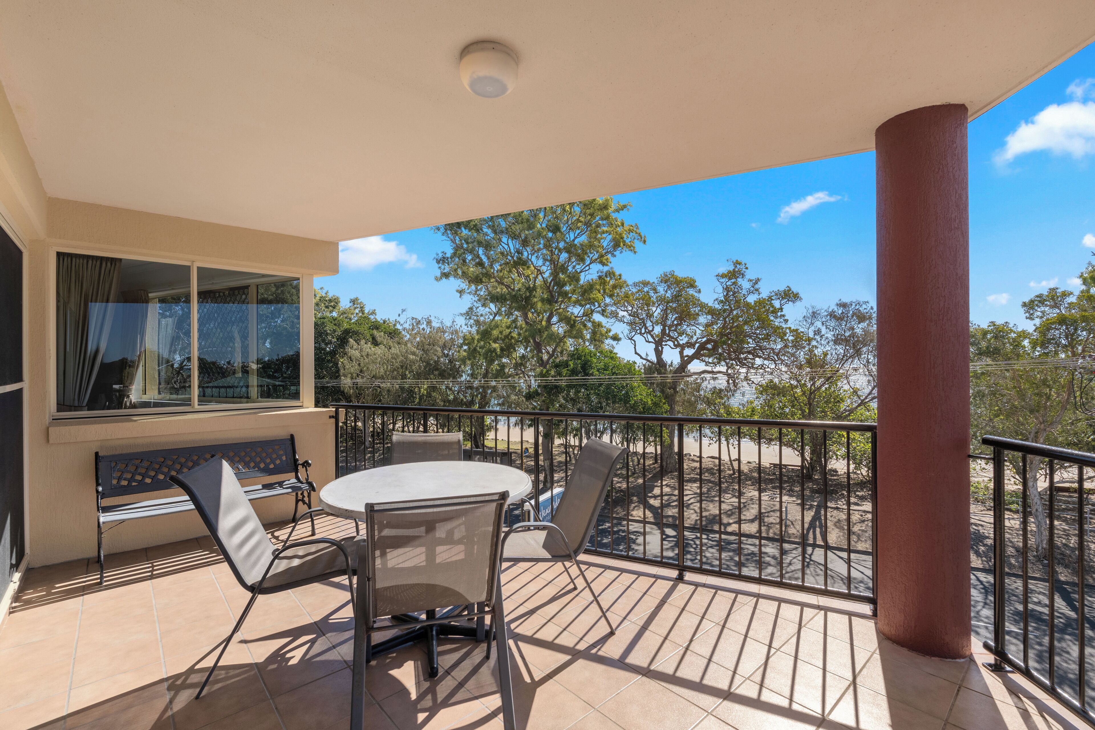 A balcony with a table and chairs, a bench, and a view of trees and a beach.