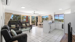 A modern living room with a sofa, coffee table, and dining area. It features a kitchen with a refrigerator, stove, and cabinets. Large windows provide ample natural light and views of the outdoors. - Alexander Beachfront Apartments (Torquay)