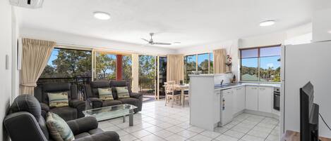 A modern living room with a sofa, coffee table, and dining area. It features a kitchen with a refrigerator, stove, and cabinets. Large windows provide ample natural light and views of the outdoors.