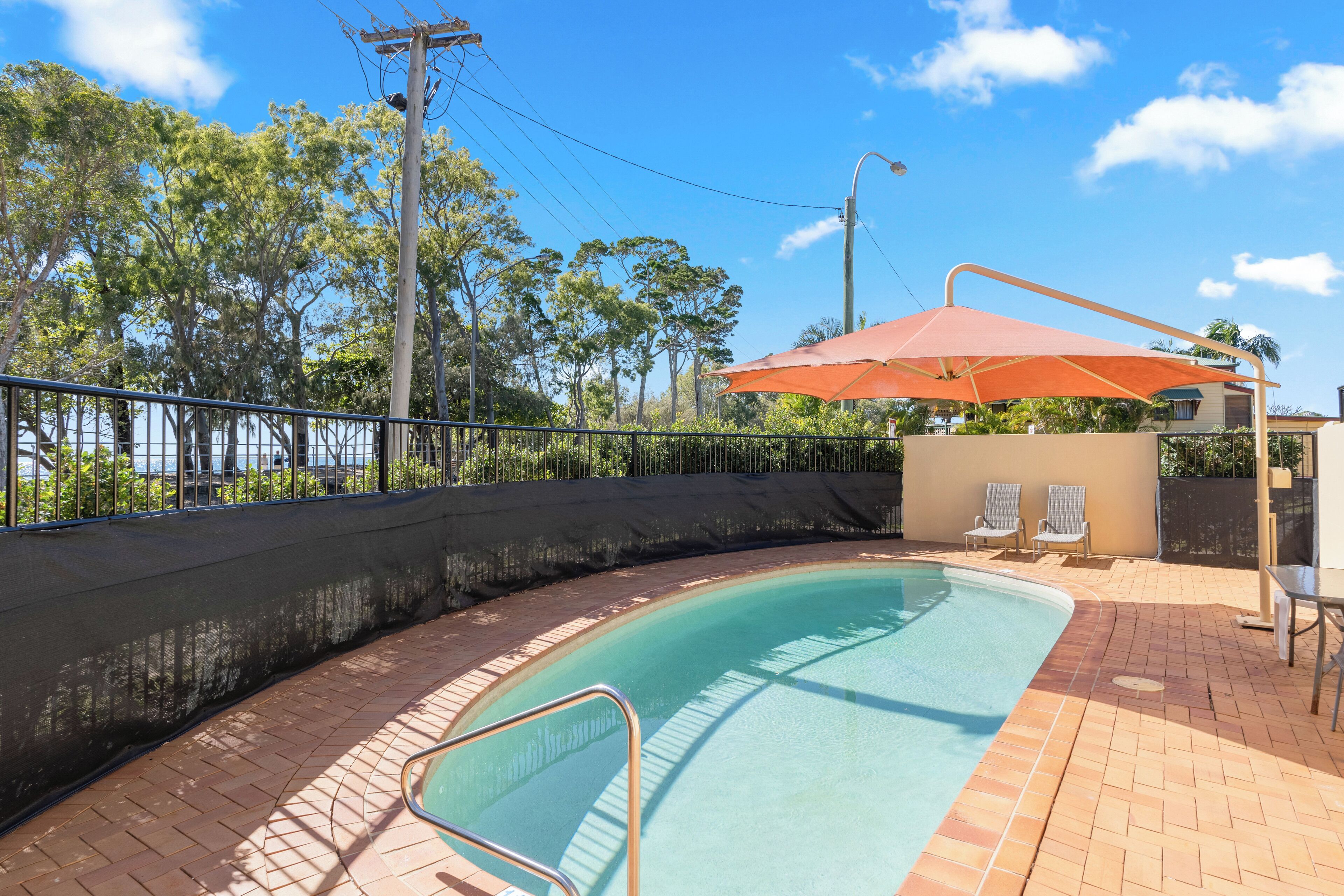 A swimming pool with a tiled deck, a metal safety railing, and a covered seating area with two chairs.