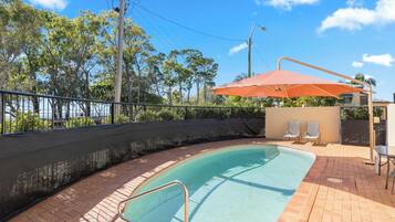 A swimming pool with a tiled deck, a metal safety railing, and a covered seating area with two chairs.