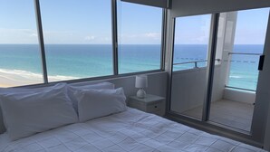 Habitación de hotel con una cama, una mesita de noche, una lámpara y vistas al océano a través de amplios ventanales.