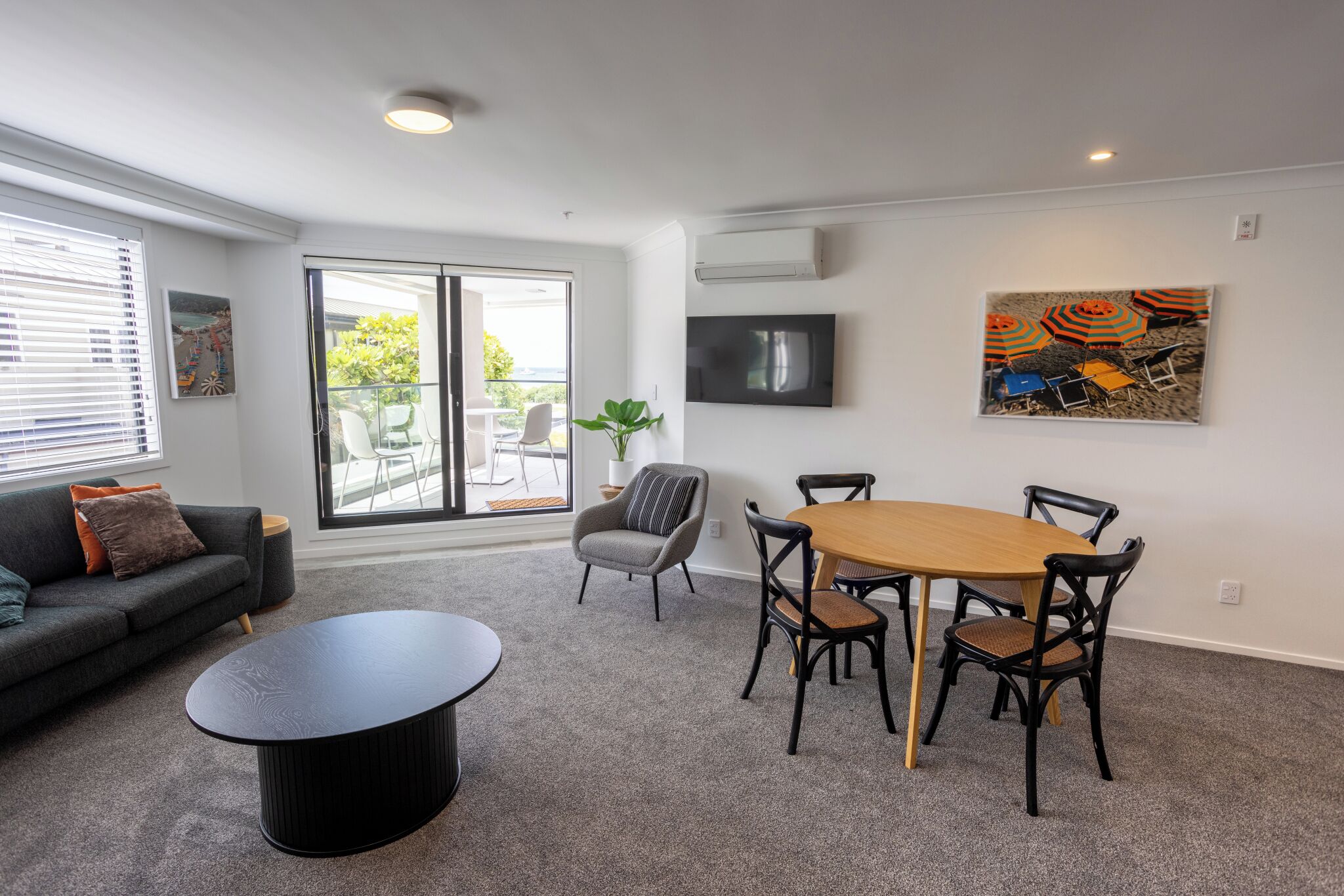 A modern living room with a sofa, a coffee table, a dining table with chairs, a TV mounted on the wall, and a sliding glass door leading to an outdoor area.