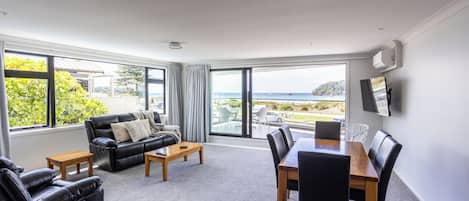 Een moderne woonkamer met een groot raam, een zwarte leren bank, een houten salontafel en een eethoek met een houten tafel en stoelen.