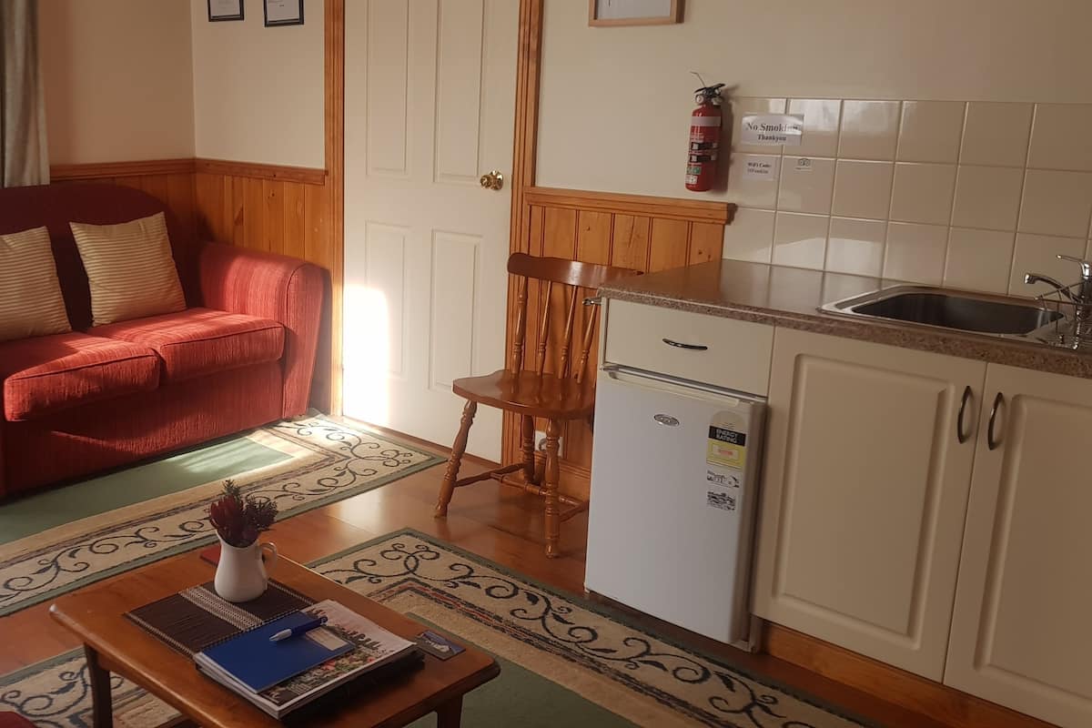 A cozy living room with a red sofa, a wooden chair, a small table with a book and a vase, and a kitchen area with a refrigerator and sink.