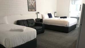 Una habitación de hotel con dos camas, un sillón de cuero oscuro, una mesita pequeña y un refrigerador blanco.