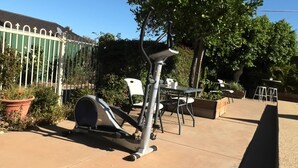 Zona de patio al aire libre con equipo de ejercicio, incluyendo una máquina elíptica y una bicicleta estática, rodeada de plantas en macetas y mobiliario de exterior.