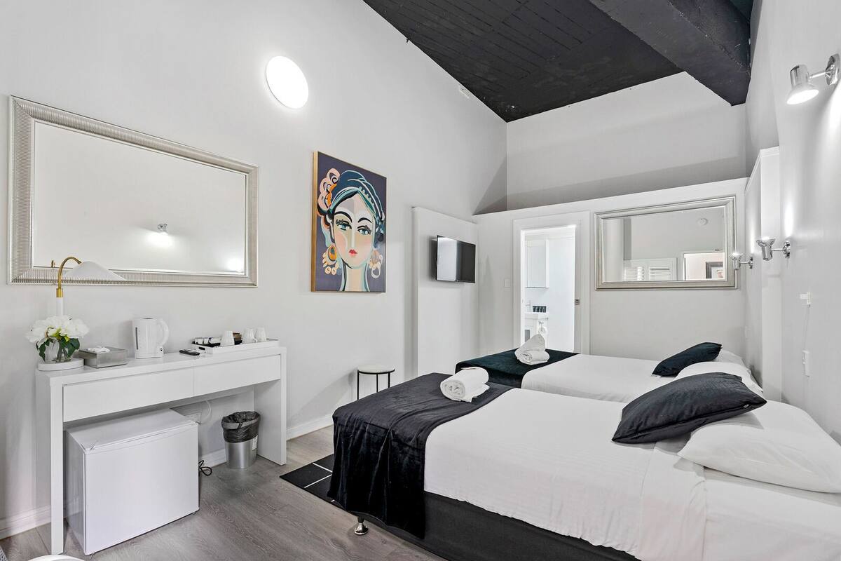 A modern hotel room with a large bed, a bedside table, a mirror, a painting, and a small kitchenette.