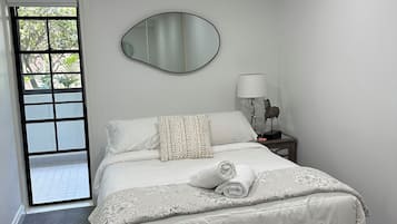 Ein Schlafzimmer mit einem Bett, einer Nachttischlampe, einem Spiegel und Blick ins Freie durch eine Tür.