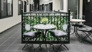 Uma pequena área externa com espaço para sentar, com uma mesa redonda e quatro cadeiras, cercada por um portão de metal decorativo.