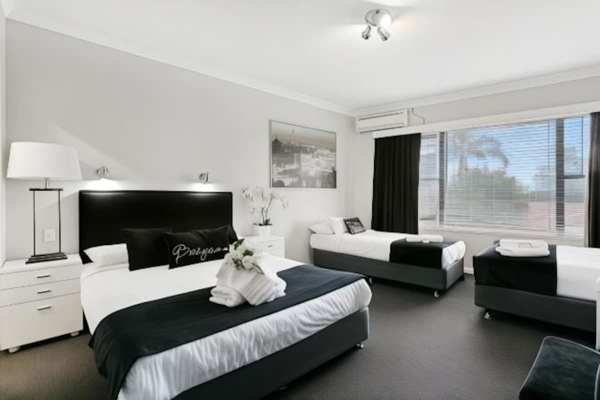 A modern hotel room with a large bed, two smaller beds, a desk, and a window with curtains.
