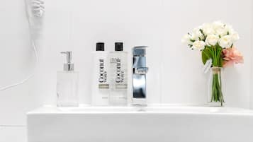 Une salle de bain avec un meuble-lavabo blanc, un sèche-cheveux, deux bouteilles d’eau de coco, un robinet et un vase avec des fleurs.
