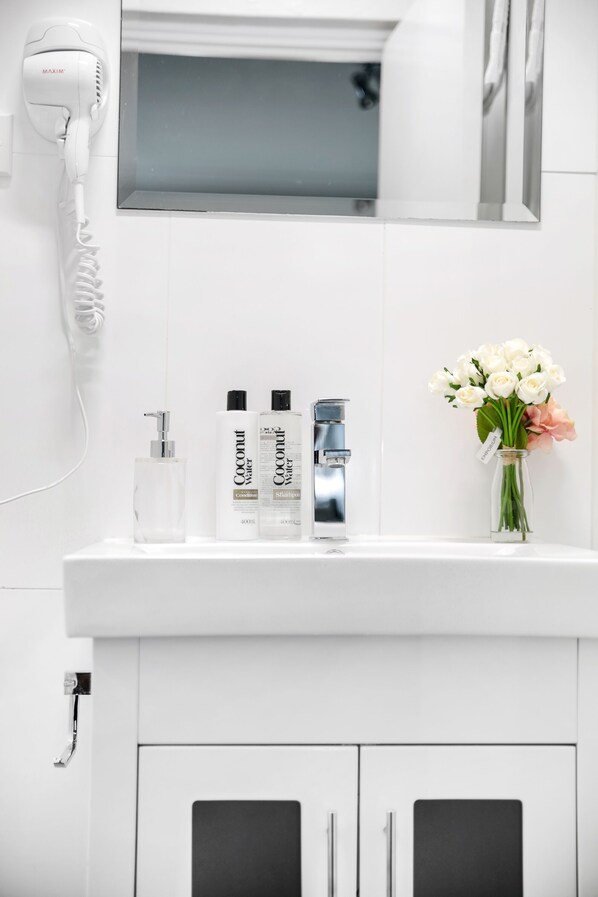 Casa de banho com toucador branco, secador de cabelo, duas garrafas de água de coco, torneira e um vaso com flores.