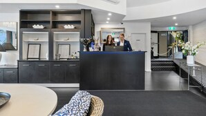 Uma área de recepção moderna com um balcão escuro, prateleiras com objetos decorativos e um espaço de estar com uma mesa redonda e almofadas estampadas.