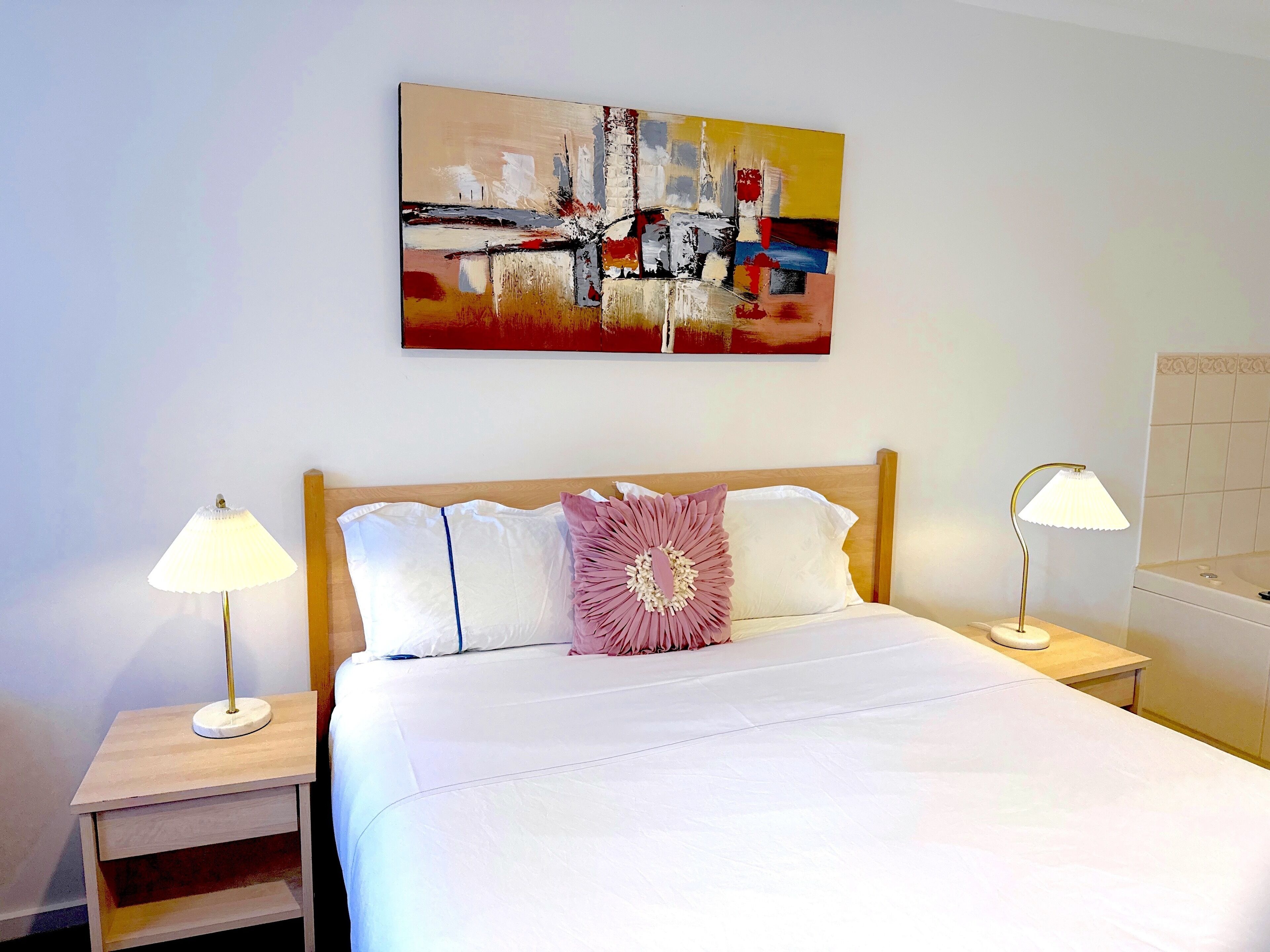 A hotel room with a bed, two bedside tables, a nightstand, a wall-mounted painting, and a bathroom with a bathtub.