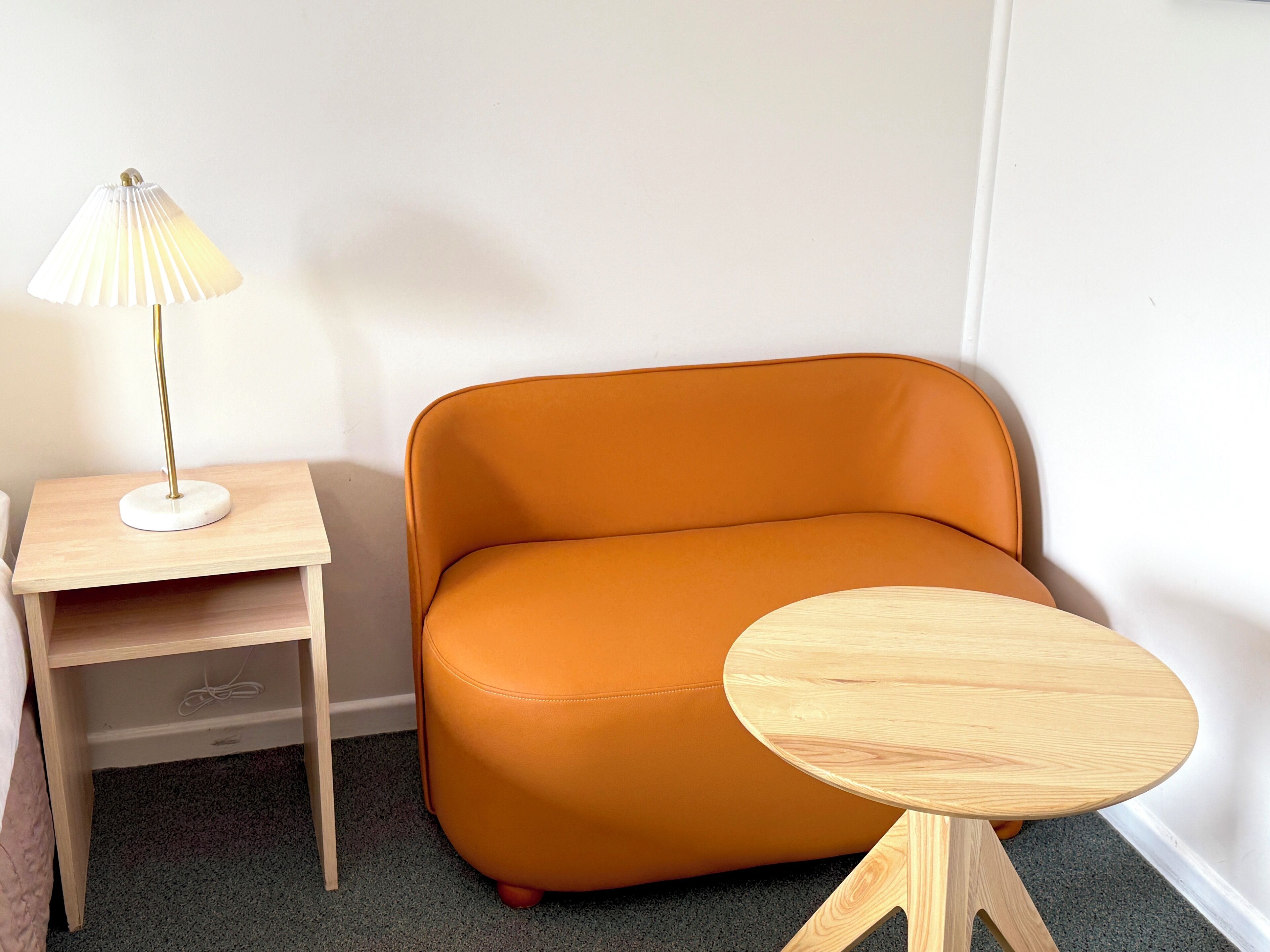 Habitación de esquina con mesita de noche de madera, una pequeña mesa y un sillón de cuero naranja.