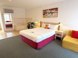 Una habitación de hotel con una cama grande, una mesita de noche con lámpara, otra mesita de noche más pequeña también con lámpara, un sofá, una planta y un cuadro en la pared.