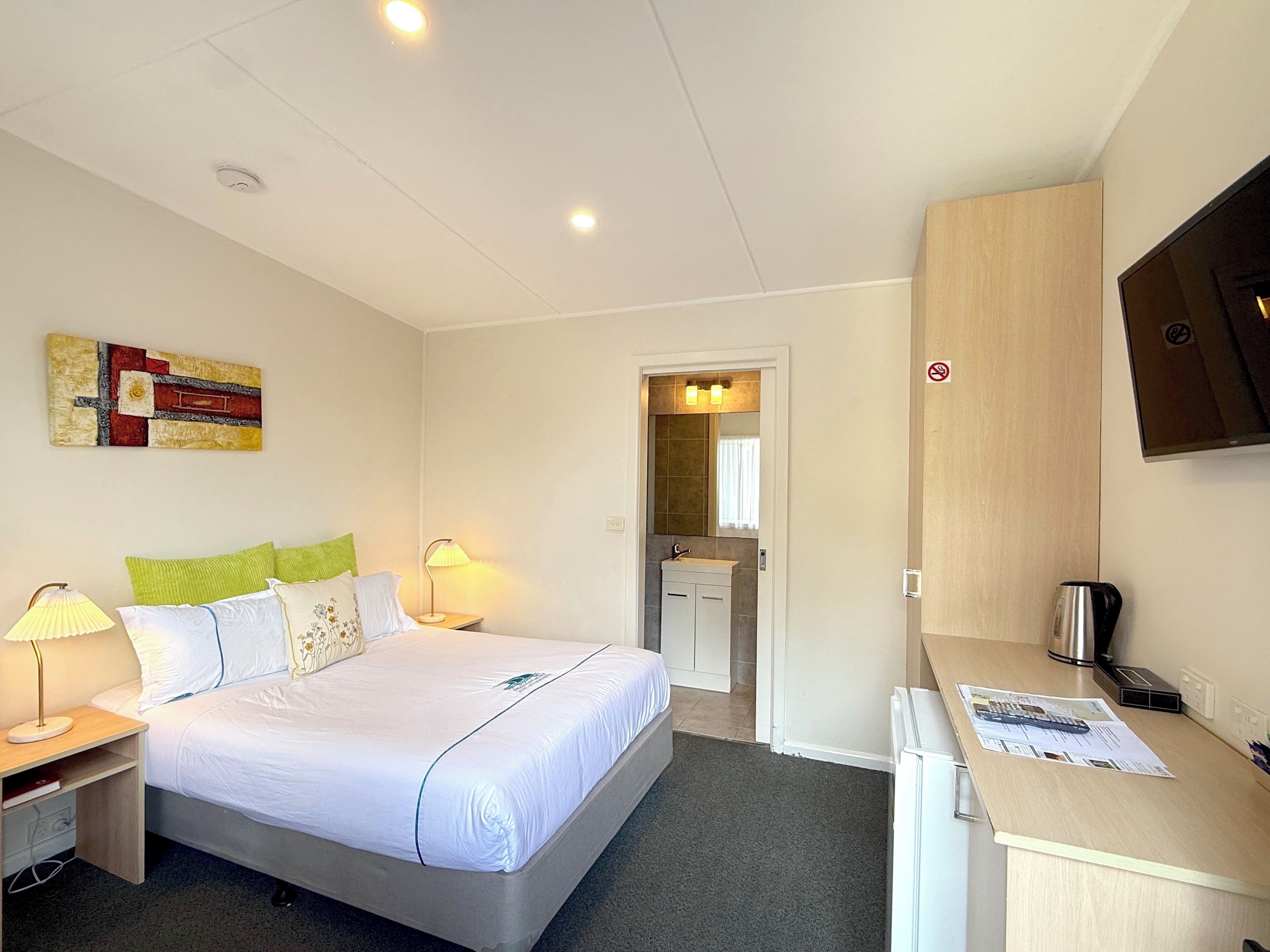 Une chambre d’hôtel avec un lit, des lampes de chevet, un bureau avec un ordinateur, un téléviseur fixé au mur et une salle de bain visible à travers une porte ouverte.