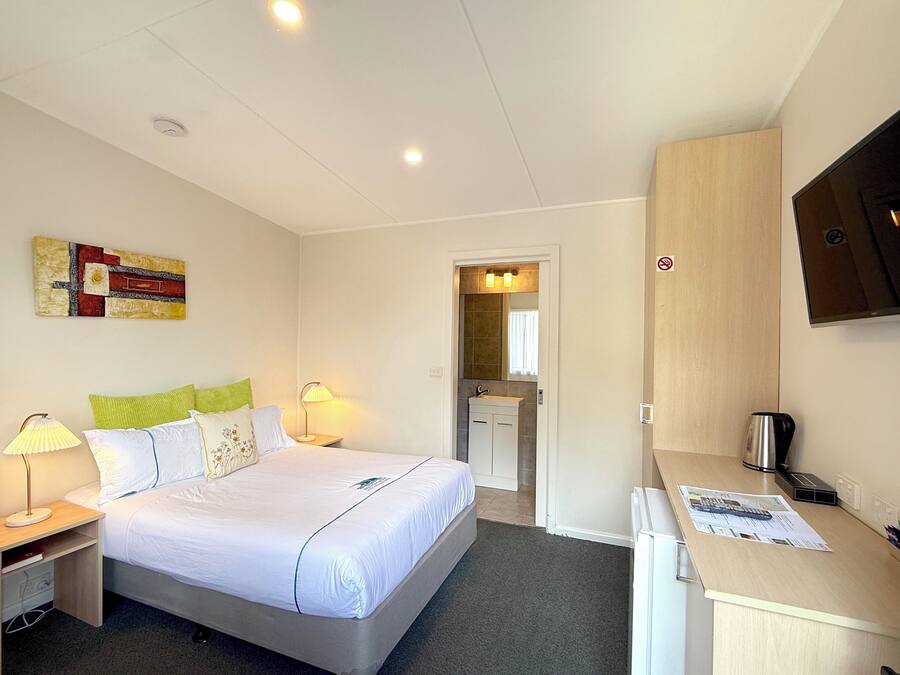 Une chambre d’hôtel avec un lit, des lampes de chevet, un bureau avec un ordinateur, un téléviseur fixé au mur et une salle de bain visible à travers une porte ouverte.