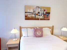 A neatly made bed with white bedding and a decorative pillow, flanked by bedside tables with lamps, in a room with a large abstract painting on the wall.