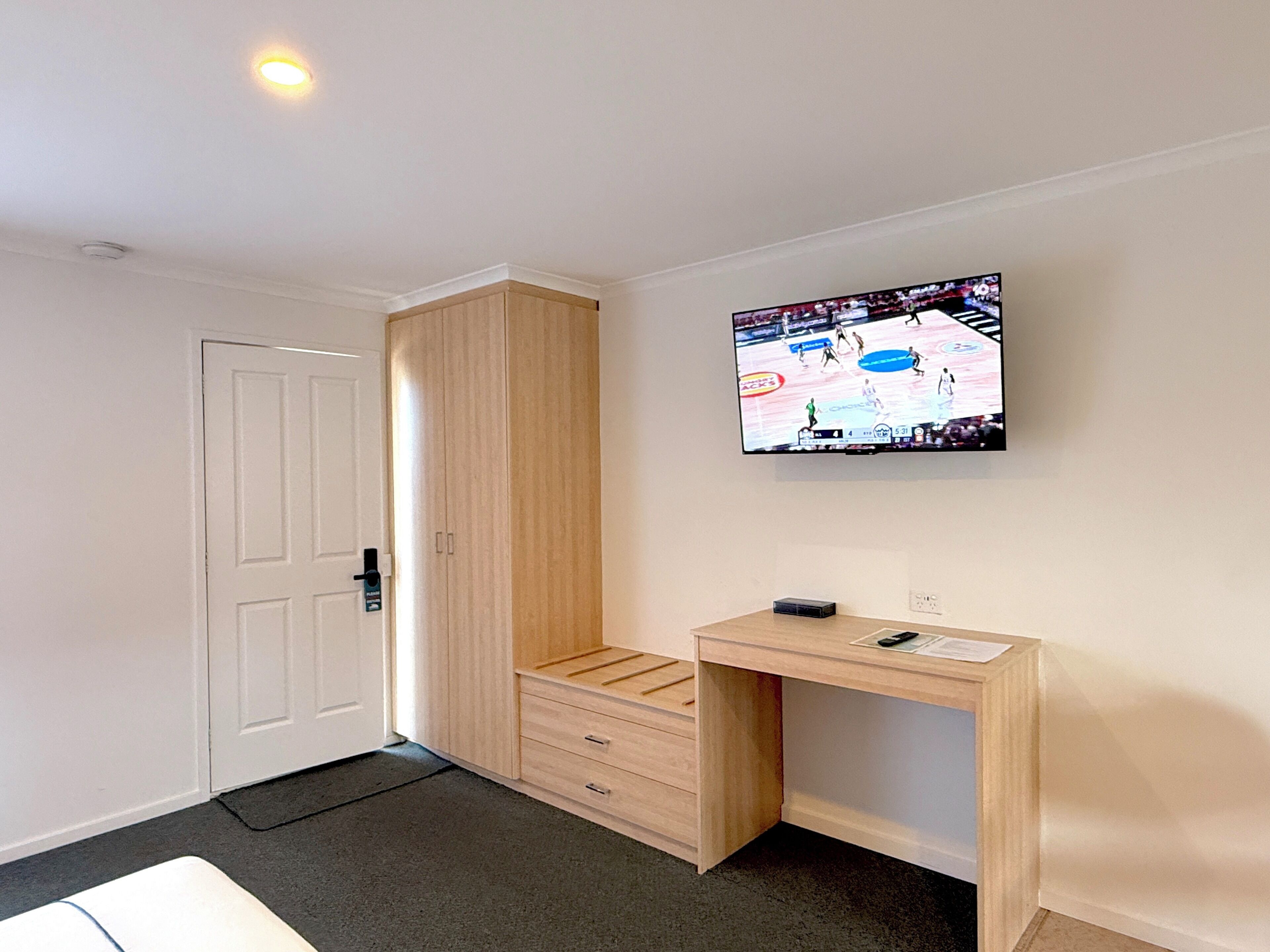 A room with a wooden wardrobe, a desk with a chair, and a television mounted on the wall displaying a sports event.