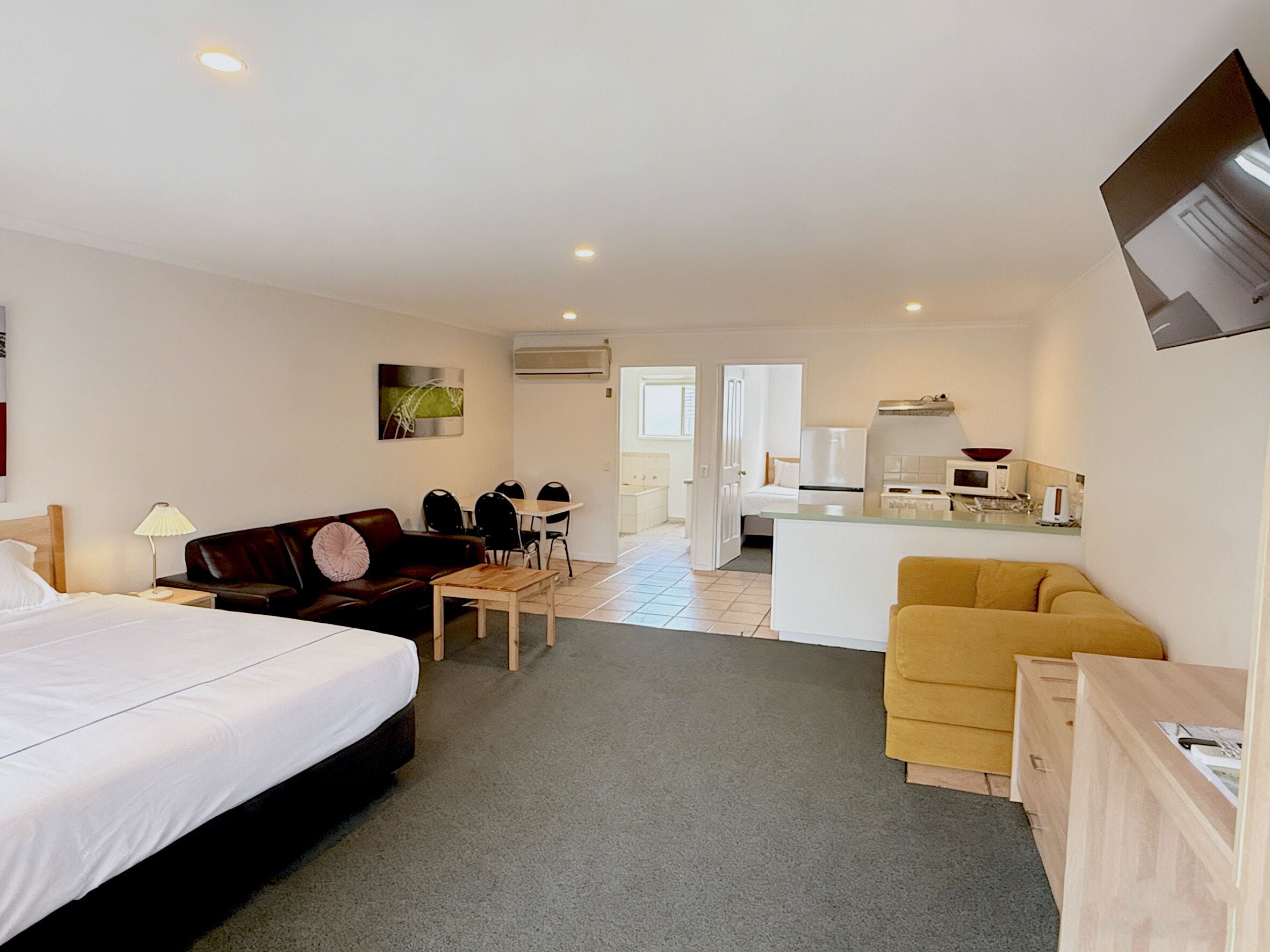 A hotel room with a bed, a sofa, a dining table with chairs, a kitchen area with a microwave, and a small desk.