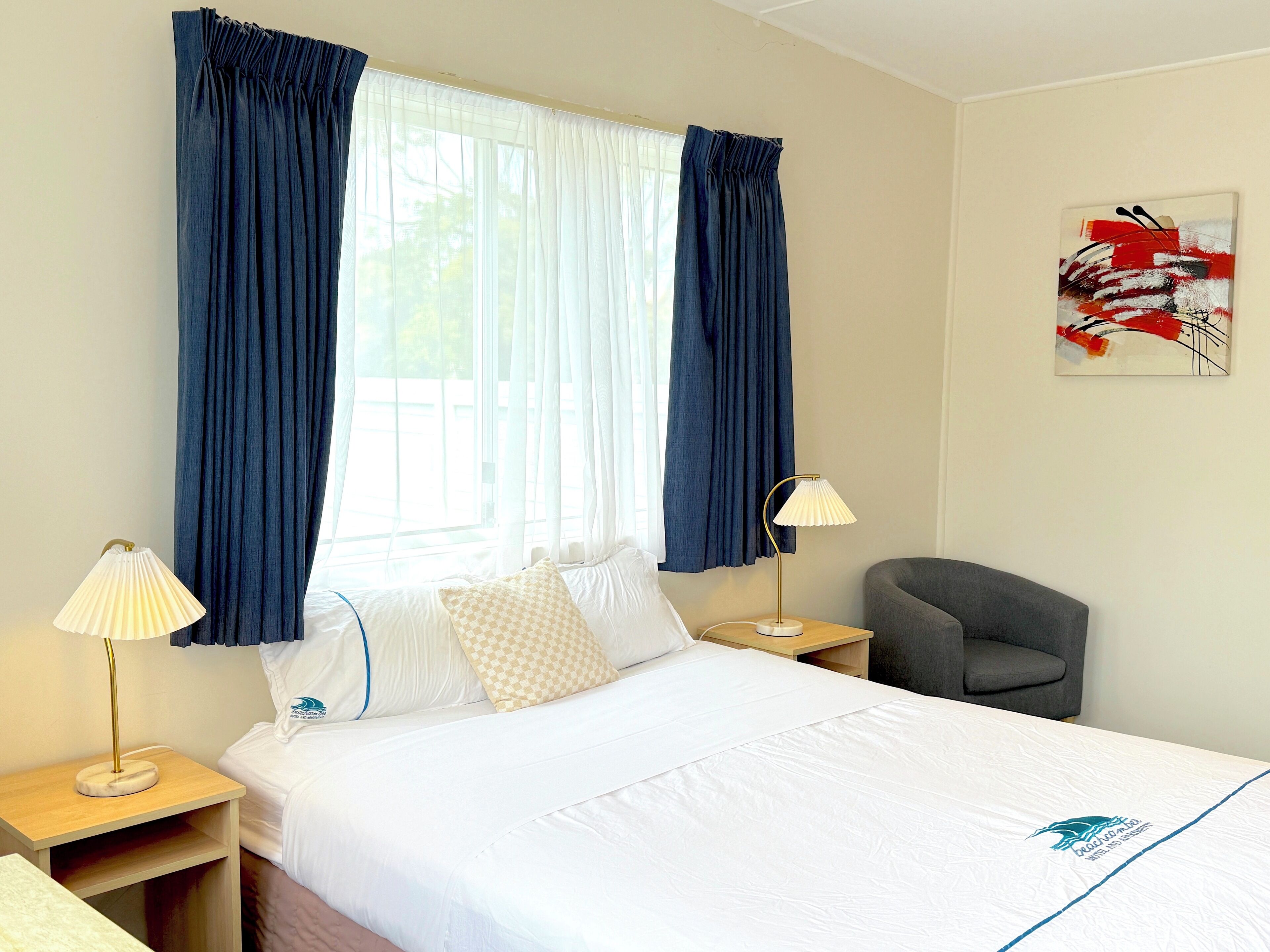 Une chambre d’hôtel avec un lit, deux lampes de chevet, une chaise, une fenêtre avec des rideaux et un tableau au mur.
