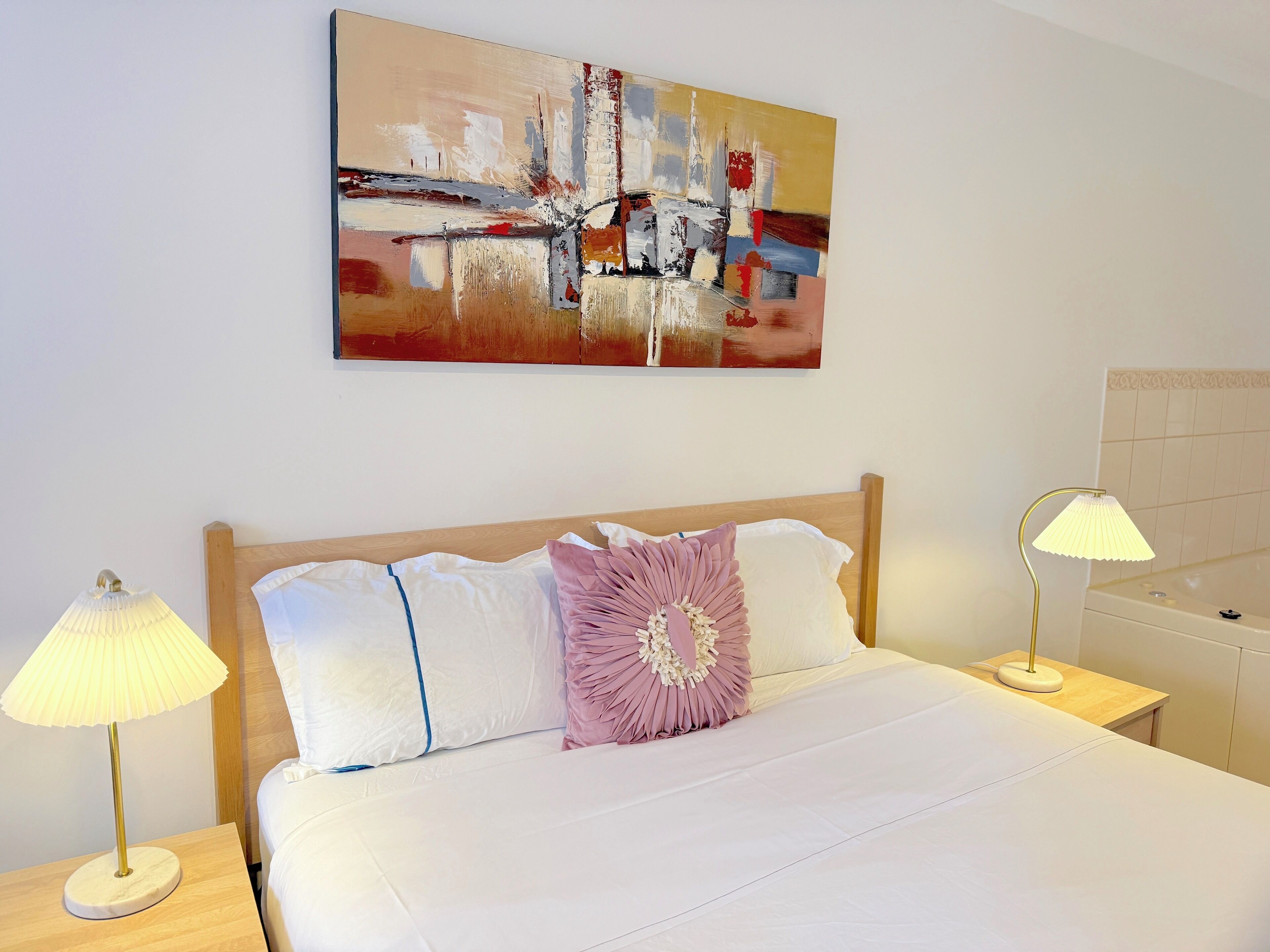 A neatly made bed with white and pink pillows, two bedside lamps, and a modern abstract painting on the wall.