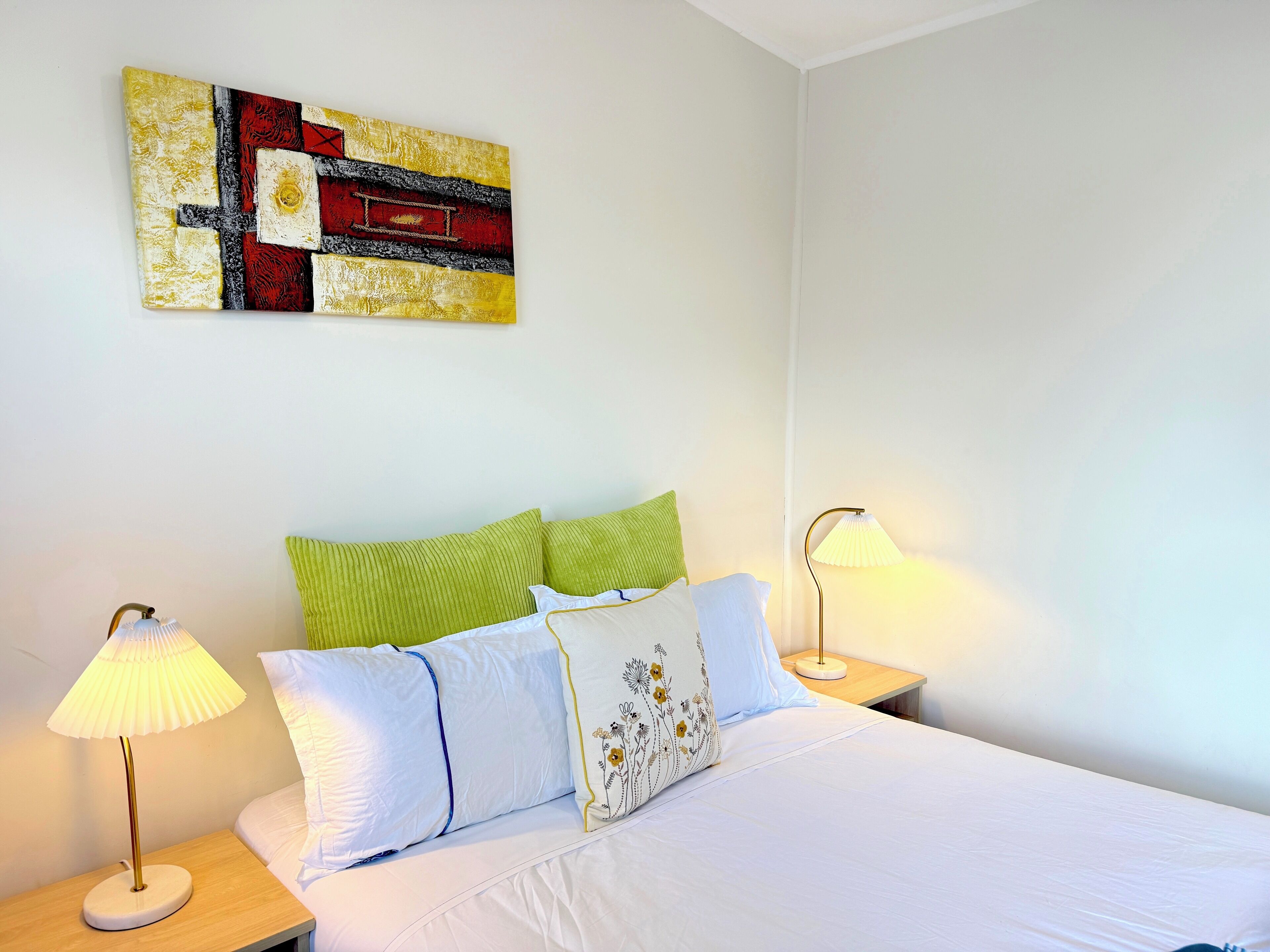 Een net opgemaakt bed met wit beddengoed en decoratieve kussens, een modern abstract schilderij erboven en twee bedlampen op houten nachtkastjes.