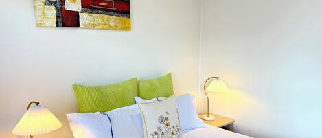 Een net opgemaakt bed met wit beddengoed en decoratieve kussens, een modern abstract schilderij erboven en twee bedlampen op houten nachtkastjes.
