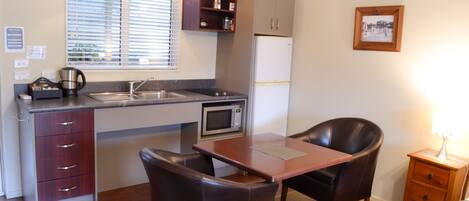 A compact kitchen with a microwave, sink, and cabinets. There is a dining table with chairs and a small bedside table with a lamp.