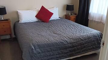 A neatly made bed with a grey quilt, a red pillow, and two bedside tables with lamps.