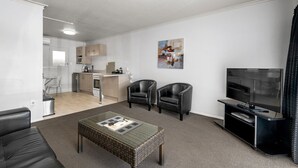 Una sala moderna con televisor, dos sillones de cuero, una mesa de centro y una cocina al fondo.