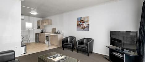 A modern living room with a TV, two leather chairs, a coffee table, and a kitchen in the background.