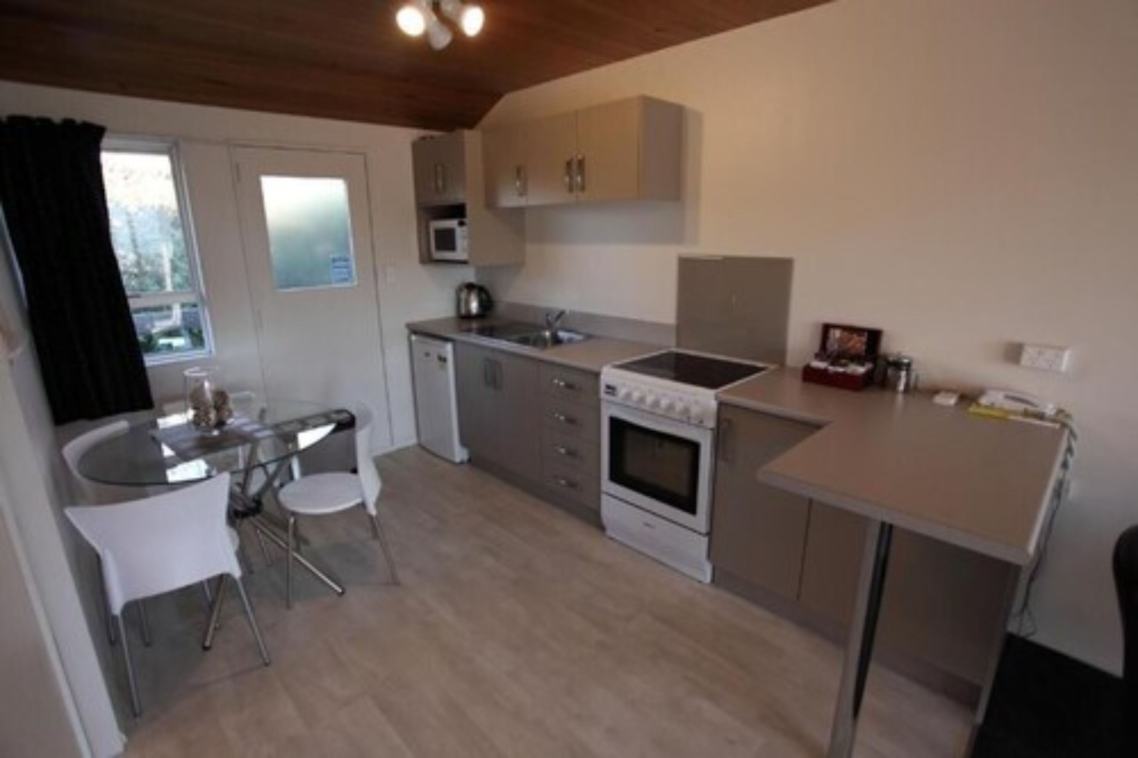 Una cocina compacta con microondas, estufa, lavaplatos y un pequeño comedor con una mesa de vidrio y sillas blancas.